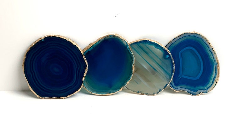 May include: Four blue agate coasters with gold gilded edges are arranged in a row against a white background. The coasters vary in shades of blue, from deep navy to light teal, and have unique natural patterns.