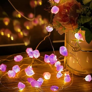 May include: Purple and clear crystal shaped fairy lights strung on a copper wire. The lights are strung around a wooden table and a vase of pink flowers.