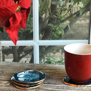 May include: A red ceramic mug and a stack of four blue and gold agate coasters sit on a weathered wooden surface. A single coaster is under the mug. A glass jar with red poinsettia leaves is in the background.