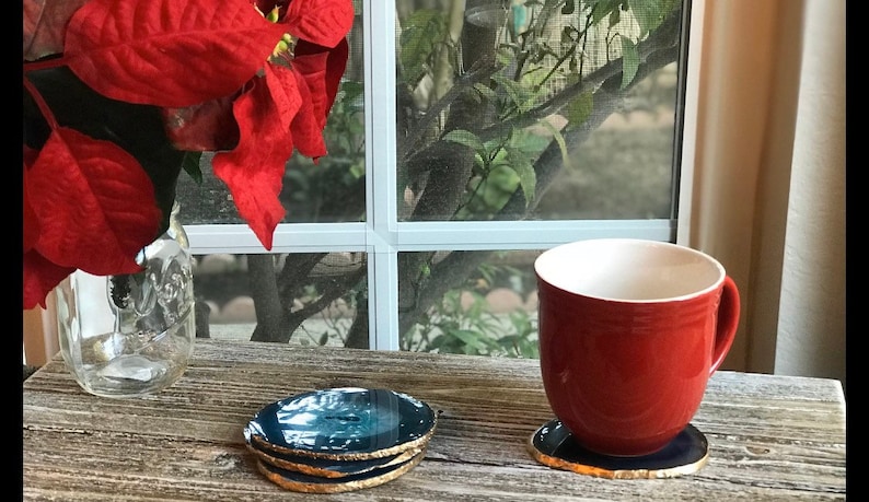 May include: A red ceramic mug and a stack of four blue and gold agate coasters sit on a weathered wooden surface. A single coaster is under the mug. A glass jar with red poinsettia leaves is in the background.