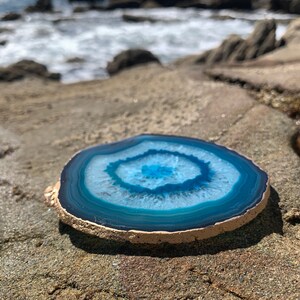 May include: A round, polished agate slice with concentric bands of blue and white. The stone has a natural, rough edge and is displayed on a textured, light brown surface, with the ocean in the background.