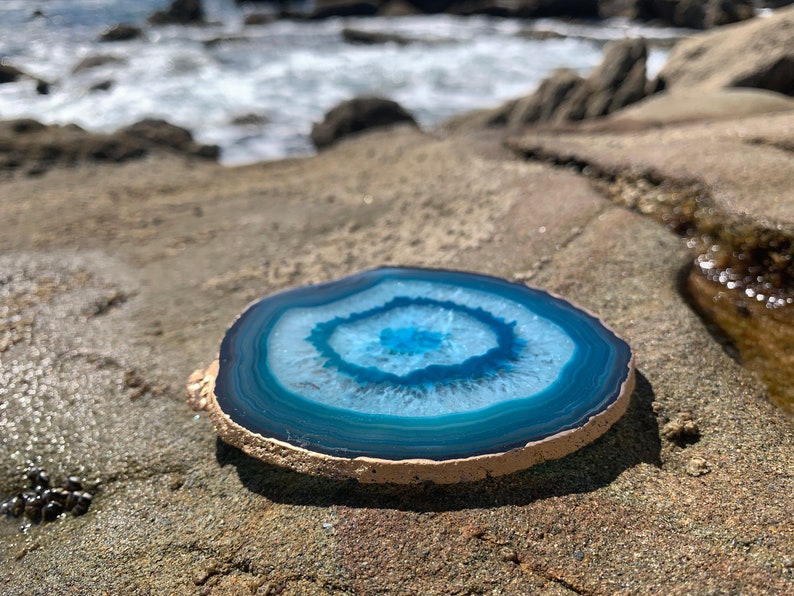 May include: A round, polished agate slice with concentric bands of blue and white. The stone has a natural, rough edge and is displayed on a textured, light brown surface, with the ocean in the background.