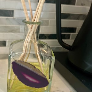May include: A clear glass bottle with a square base filled with a yellow liquid and a purple agate slice. The bottle has wooden sticks inserted into it, which are used to diffuse the scent of the liquid.