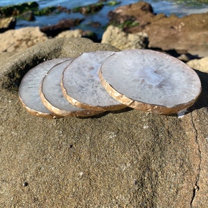 以下が含まれることがあります： 岩の表面に積み重ねられた、金色の縁取りが施された白い瑪瑙のコースター4枚