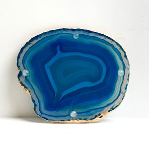 May include: A polished, natural agate slice in shades of blue and white, with a gold-painted edge. Four small, clear, round, decorative accents are evenly spaced on the surface. The stone's layered pattern is visible.