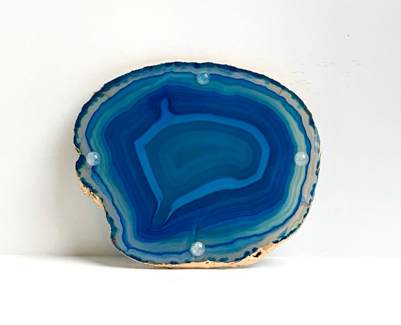 May include: A polished, natural agate slice in shades of blue and white, with a gold-painted edge. Four small, clear, round, decorative accents are evenly spaced on the surface. The stone's layered pattern is visible.