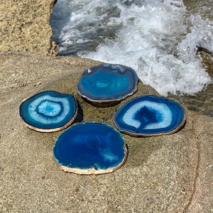 May include: Four blue agate coasters with gold-painted edges are arranged on a rock. The coasters have varying shades of blue and white patterns. The background shows a body of water with white waves.