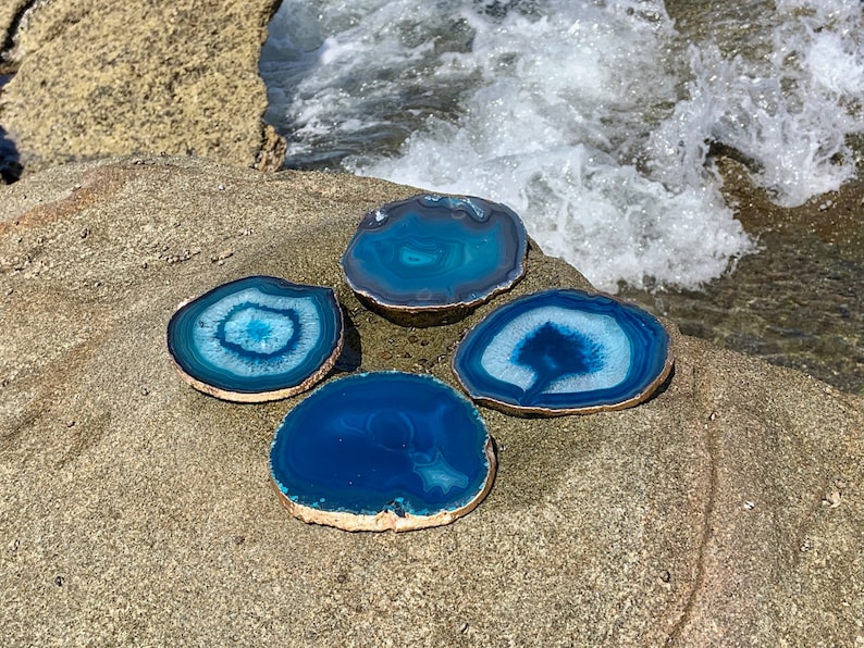 May include: Four blue agate coasters with gold-painted edges are arranged on a rock. The coasters have varying shades of blue and white patterns. The background shows a body of water with white waves.
