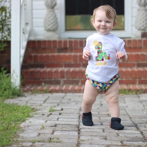 May include: A young child wearing a white t-shirt with a colorful number one and a caterpillar design. The shirt says "Jack" on it. The child is wearing colorful patterned diaper shorts and black shoes. The child is standing on a brick walkway.