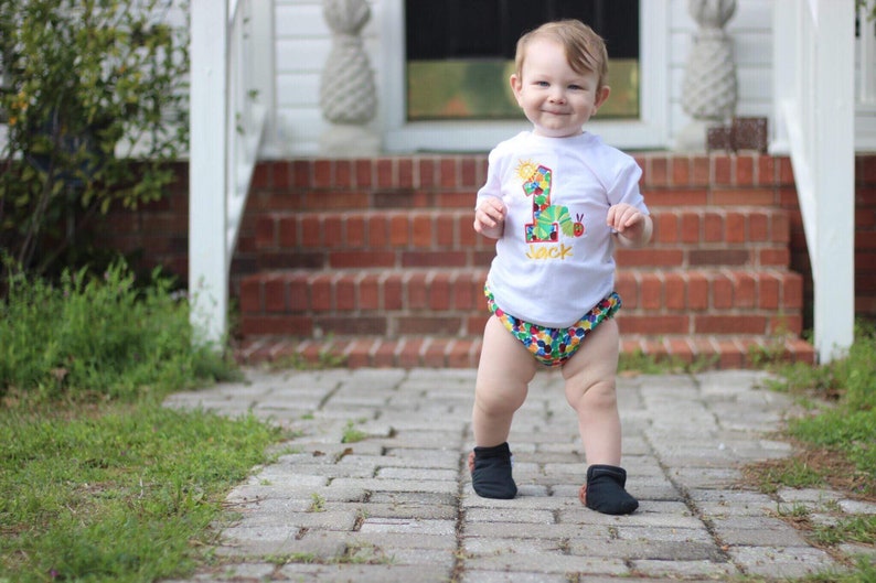May include: A young child wearing a white t-shirt with a colorful number one and a caterpillar design. The shirt says "Jack" on it. The child is wearing colorful patterned diaper shorts and black shoes. The child is standing on a brick walkway.