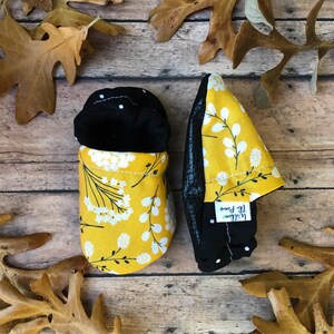 May include: A pair of yellow and black baby booties with a white floral pattern. The booties have a black leather sole and a white label that says "Little Feet" on them.