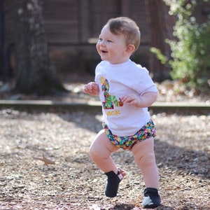 May include: A toddler wearing a white t-shirt with a colorful caterpillar graphic and the text "One" on it. The toddler is wearing colorful patterned bloomers and black shoes. The toddler is standing on a brown gravel surface.