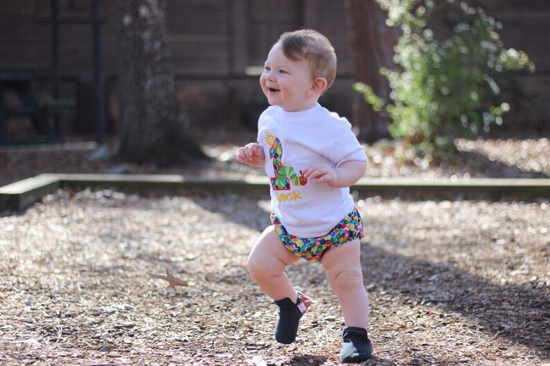 May include: A toddler wearing a white t-shirt with a colorful caterpillar graphic and the text "One" on it. The toddler is wearing colorful patterned bloomers and black shoes. The toddler is standing on a brown gravel surface.