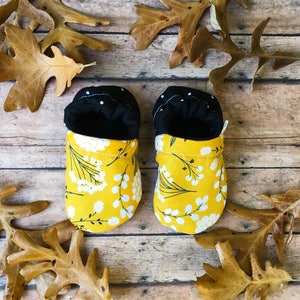 May include: A pair of yellow baby booties with a white floral pattern. The booties have black fabric lining and a white polka dot pattern on the lining.