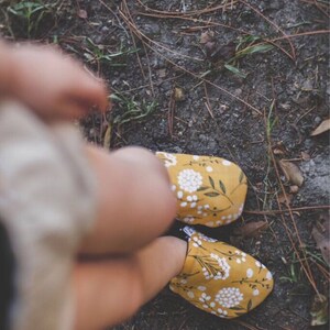May include: A pair of yellow fabric slippers with a white floral pattern. The slippers are worn by a person standing on a dirt path.