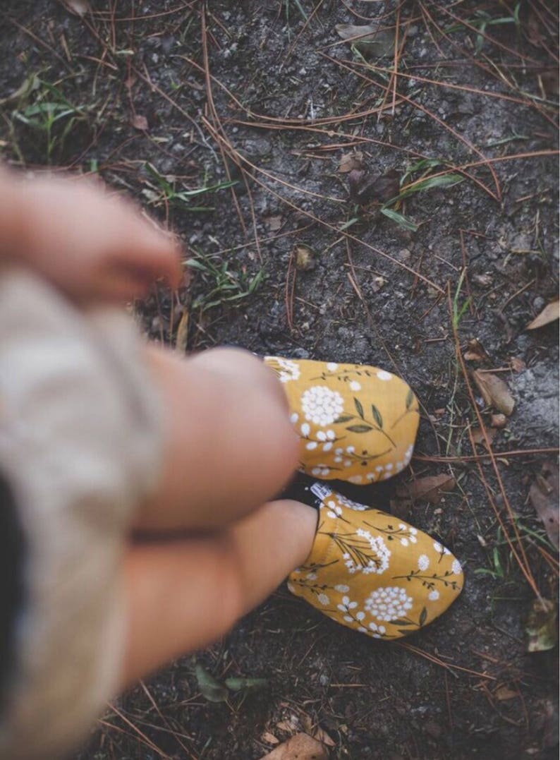 May include: A pair of yellow fabric slippers with a white floral pattern. The slippers are worn by a person standing on a dirt path.