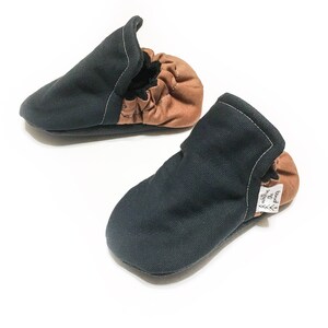 May include: A pair of black and brown baby shoes with a soft fabric upper and a flexible sole. The shoes have a label that says "The Willow Tree".