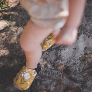 May include: A pair of yellow and black floral print shoes with black soles. The shoes are worn by a child standing on a dirt path.