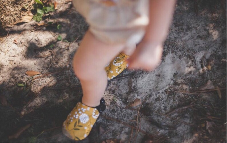 May include: A pair of yellow and black floral print shoes with black soles. The shoes are worn by a child standing on a dirt path.