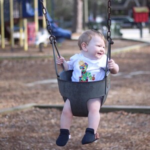 May include: A baby wearing a white shirt with a colorful number one and a caterpillar design is sitting on a swing set at a playground. The baby is smiling and looking to the right.