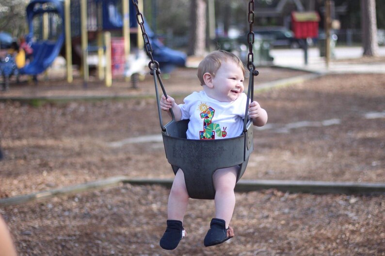May include: A baby wearing a white shirt with a colorful number one and a caterpillar design is sitting on a swing set at a playground. The baby is smiling and looking to the right.
