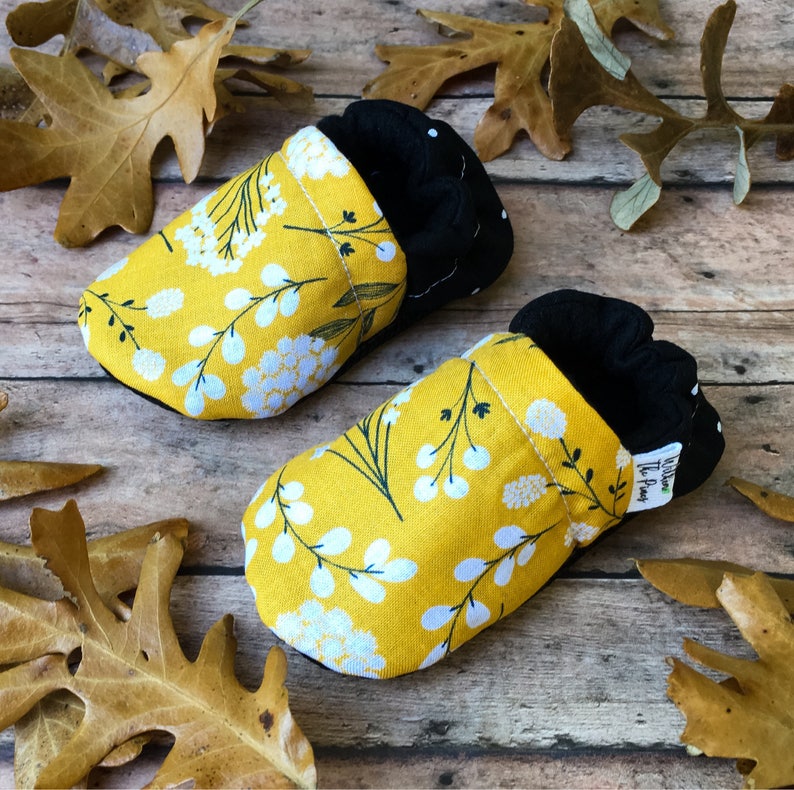 May include: A pair of yellow baby booties with a white floral pattern. The booties have black fabric on the inside and a black polka dot pattern on the back.