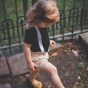 May include: A young child wearing a black short-sleeved shirt with white suspenders and beige shorts. The child is wearing yellow floral shoes and is walking on a path with green grass and brown rocks.