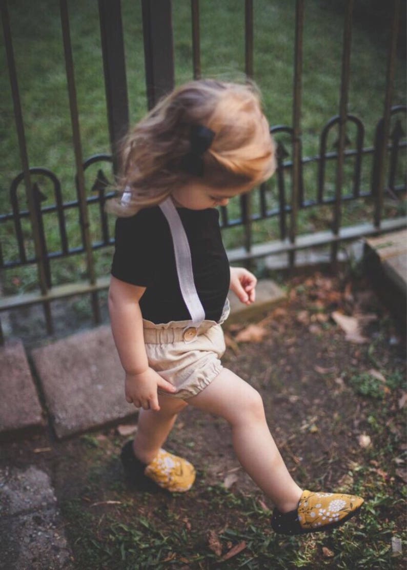 May include: A young child wearing a black short-sleeved shirt with white suspenders and beige shorts. The child is wearing yellow floral shoes and is walking on a path with green grass and brown rocks.
