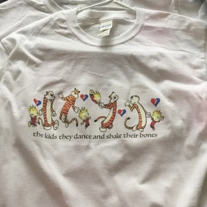 May include: White t-shirt featuring a cartoon graphic of dancing tigers and a boy, with the text "the kids they dance and shake their bones". The design includes orange, red, blue, and yellow colors.