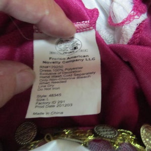 May include: A pink dress with gold trim and a white label with the text "Franco American Novelty Company LLC" and care instructions. The label also includes the style number 48345, size L, factory ID 291, and production date 201203. The dress is made in China.