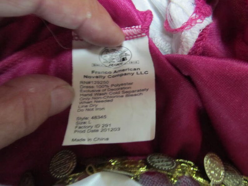 May include: A pink dress with gold trim and a white label with the text "Franco American Novelty Company LLC" and care instructions. The label also includes the style number 48345, size L, factory ID 291, and production date 201203. The dress is made in China.