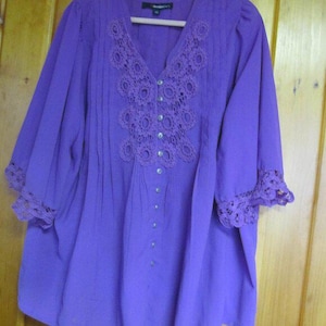 May include: A vivid purple blouse with a V-neck and three-quarter sleeves. The blouse has detailed lace around the neckline and cuffs, and small buttons down the front. The fabric looks soft and flowing. The blouse is approximately 76 cm long.