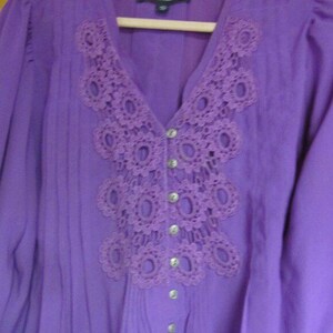 May include: A purple blouse with a V-neck and long sleeves. The neckline features a decorative, openwork lace design. The blouse has a button-down front with silver-coloured buttons. The fabric has vertical pleats.