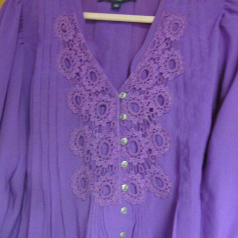 May include: A purple blouse with a V-neck and long sleeves. The neckline features a decorative, openwork lace design. The blouse has a button-down front with silver-coloured buttons. The fabric has vertical pleats.
