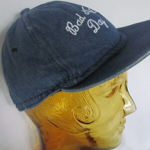 May include: A blue denim baseball cap with white embroidery that reads "Bad Day".