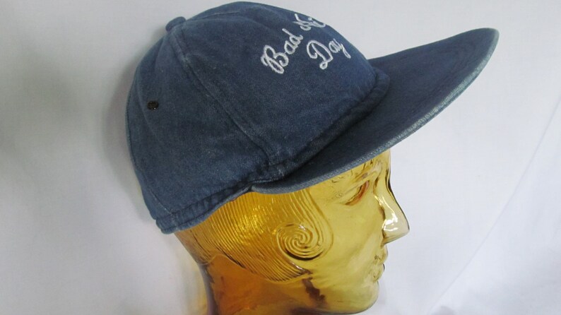 May include: A blue denim baseball cap with white embroidery that reads "Bad Day".