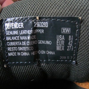 Op de afbeelding: Een zwart leren schoenetiket met witte tekst. Het etiket luidt "DEFENDER 2563293 GENUINE LEATHER UPPER BALANCE MAN MADE CUBIERTA DE CUERO GENUINO RESTO SINTETICO MADE IN CHINA HECHO EN CHINA 209 S 7" en "XW USA 8 1/2 EUR 42 MEX 27 1/2".
