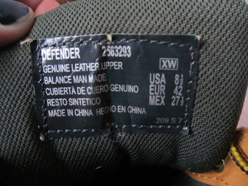 Op de afbeelding: Een zwart leren schoenetiket met witte tekst. Het etiket luidt "DEFENDER 2563293 GENUINE LEATHER UPPER BALANCE MAN MADE CUBIERTA DE CUERO GENUINO RESTO SINTETICO MADE IN CHINA HECHO EN CHINA 209 S 7" en "XW USA 8 1/2 EUR 42 MEX 27 1/2".