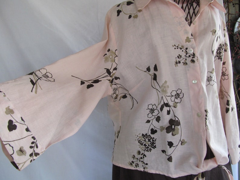 May include: A long-sleeved, light pink button-down shirt with a brown floral print. The shirt has a relaxed fit and a slightly gathered waist.