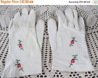 May include: A pair of white leather gloves with delicate floral embroidery in shades of green and red. The gloves are on a white crocheted surface.