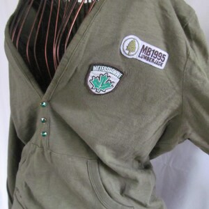 Op de afbeelding: Een groen shirt met lange mouwen, een diepe V-hals en een zak op de voorkant. Het shirt heeft drie geborduurde patches, een met een esdoornblad en de tekst "METERSBONWE", een met een boom en de tekst "MB1995" en een met de tekst "LUMBERJACK".
