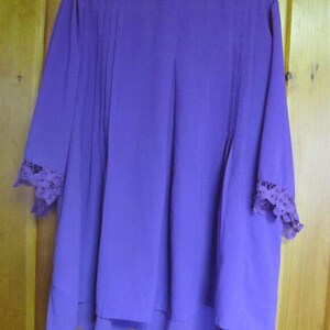 May include: A long-sleeved, purple blouse with a flowing silhouette. The blouse features vertical pleats down the front and delicate lace detailing at the cuffs. The fabric has a soft, draping quality.