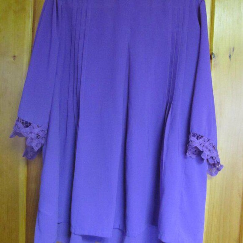 May include: A long-sleeved, purple blouse with a flowing silhouette. The blouse features vertical pleats down the front and delicate lace detailing at the cuffs. The fabric has a soft, draping quality.