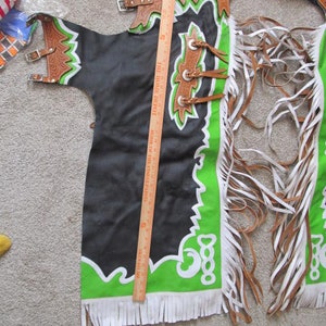 Puede incluir: Chaps de cuero negro con ribetes y flecos verdes y blancos. Las chaps tienen un dise&ntilde;o decorativo en la parte delantera y una hebilla en la parte posterior.