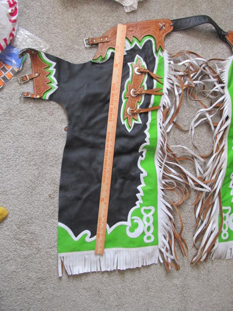 Puede incluir: Chaps de cuero negro con ribetes y flecos verdes y blancos. Las chaps tienen un dise&ntilde;o decorativo en la parte delantera y una hebilla en la parte posterior.