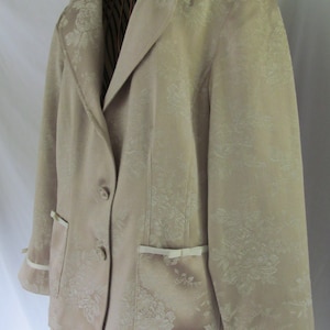 Op de afbeelding: Een beige blazer met een bloemenpatroon en witte bies. De blazer heeft een enkele knoopsluiting met twee knopen en twee zakken met witte lintaccenten.