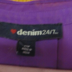 May include: Close-up of a purple garment tag with the text "denim24/7" in white, a red heart, and a smaller tag below with text. The garment is a solid purple colour.