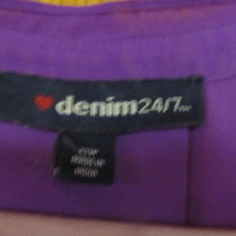 May include: Close-up of a purple garment tag with the text "denim24/7" in white, a red heart, and a smaller tag below with text. The garment is a solid purple colour.