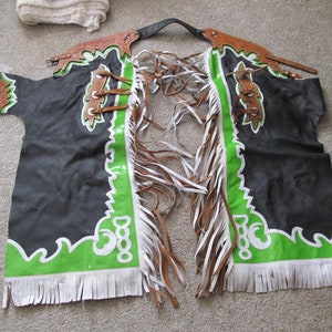 Puede incluir: Chaps de cuero negro con ribetes y flecos verdes y blancos. Las chaps tienen un dise&ntilde;o decorativo en la parte delantera.