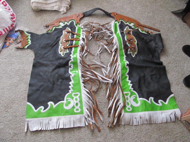 Puede incluir: Chaps de cuero negro con ribetes y flecos verdes y blancos. Las chaps tienen un dise&ntilde;o decorativo en la parte delantera.